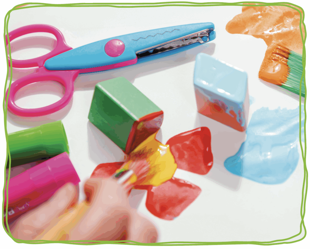 image of a child's hand painting with a paint brush and blocks with colourful scissors in the background